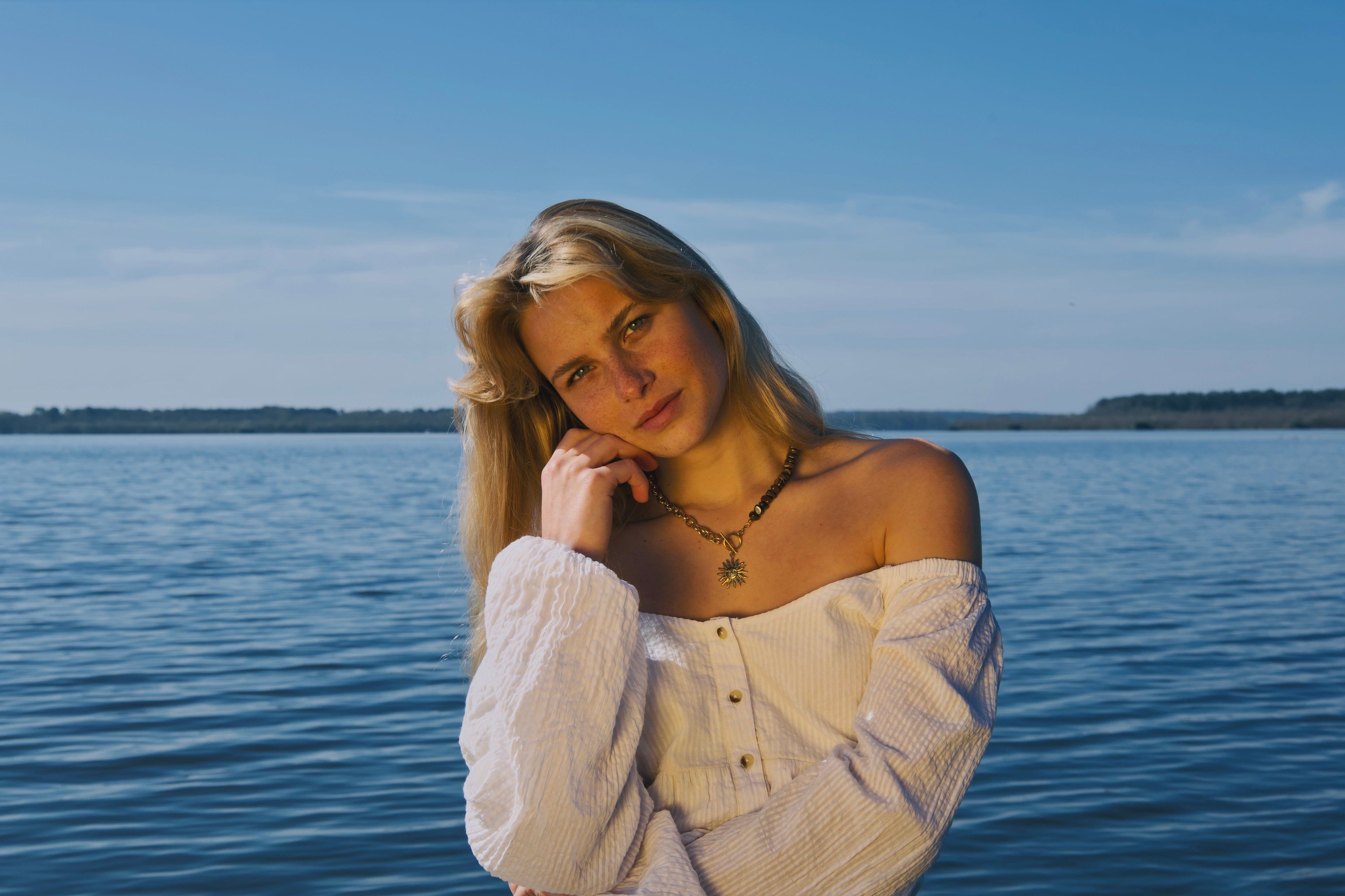 Femme devant un lac entrain de poser avec un collier fait main en pierres naturelles et acier inoxydable pour une marque de bijoux fait main